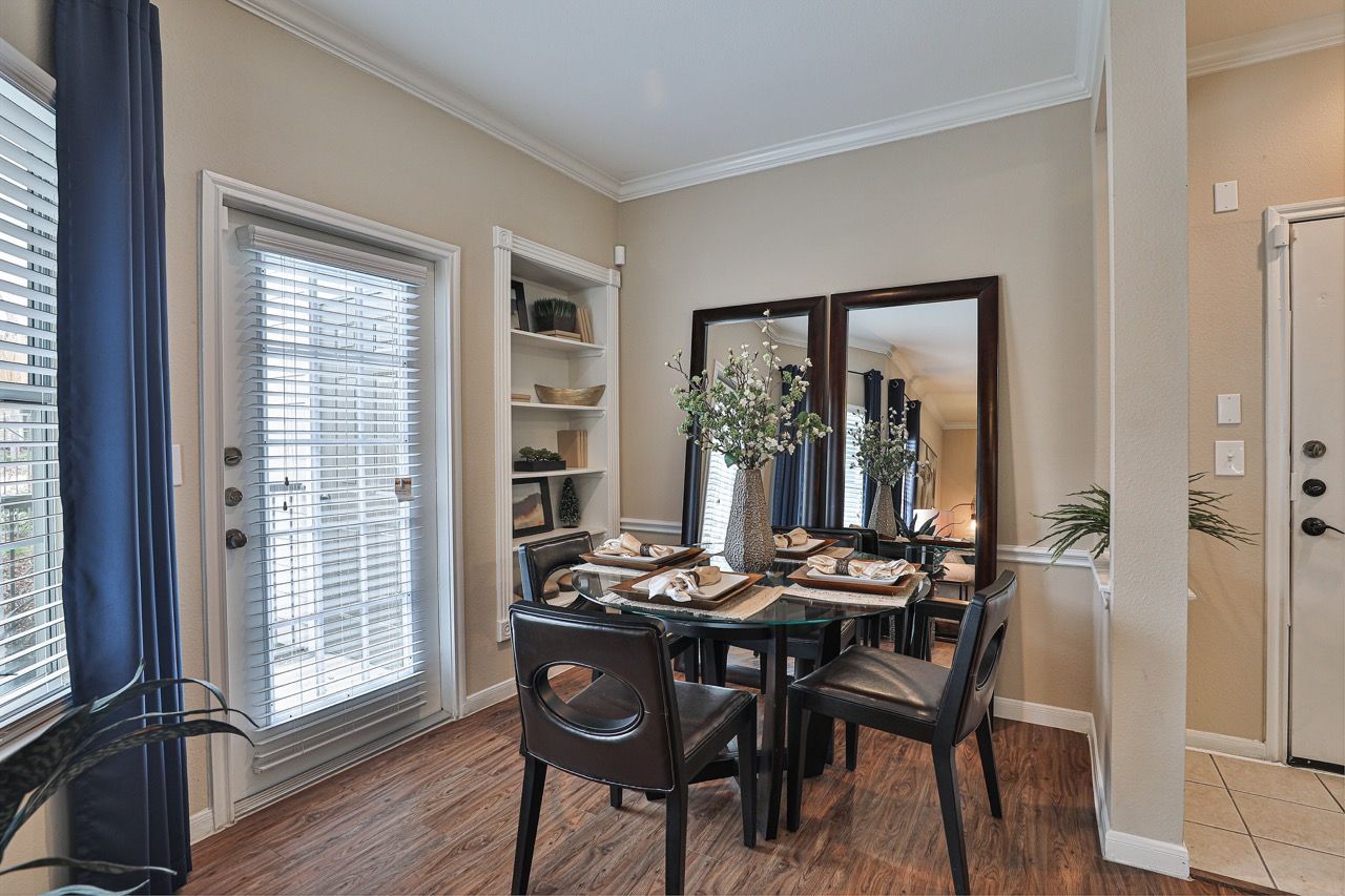Dining area in an apartment with a glass-top table, four black chairs, built-in shelves, and a large mirror.