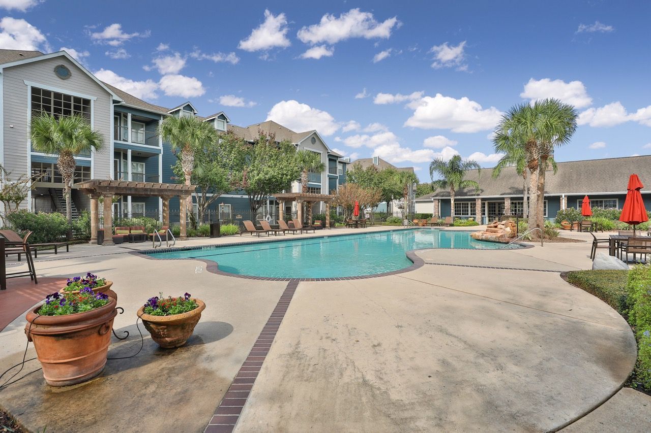 Outdoor swimming pool at an apartment complex, with patio seating, palm trees, and terracotta planters under a blue sky.