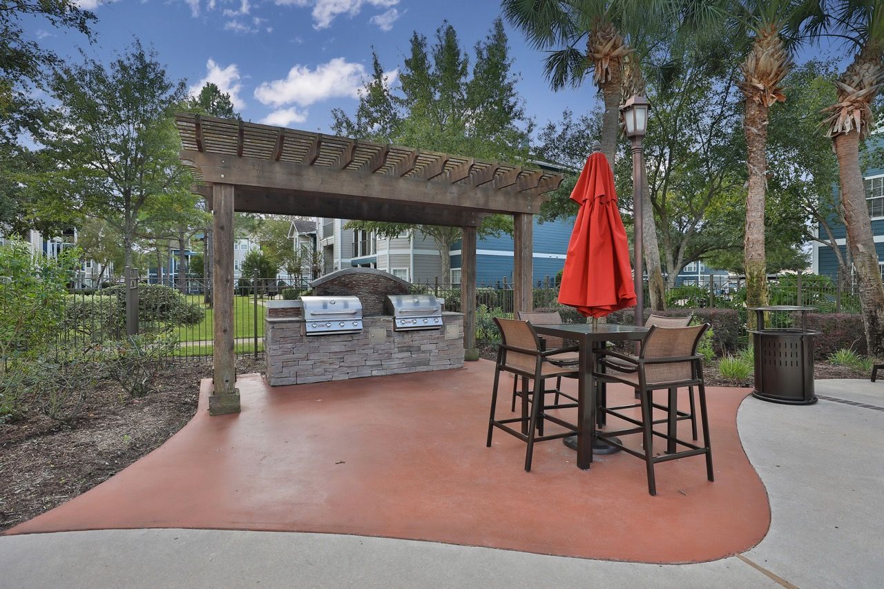 Outdoor community grilling area with a stone island, wooden pergola, and patio table with a red umbrella on a paved floor.
