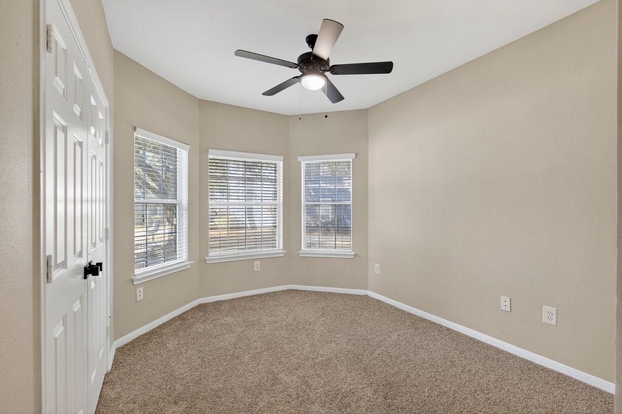Empty bedroom with beige walls, carpeted floor, three windows with blinds, and a ceiling fan.