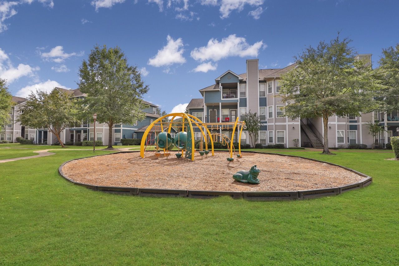 A playground with a yellow jungle gym and a small green spring rider on wood chips, set on a lawn by apartment buildings.