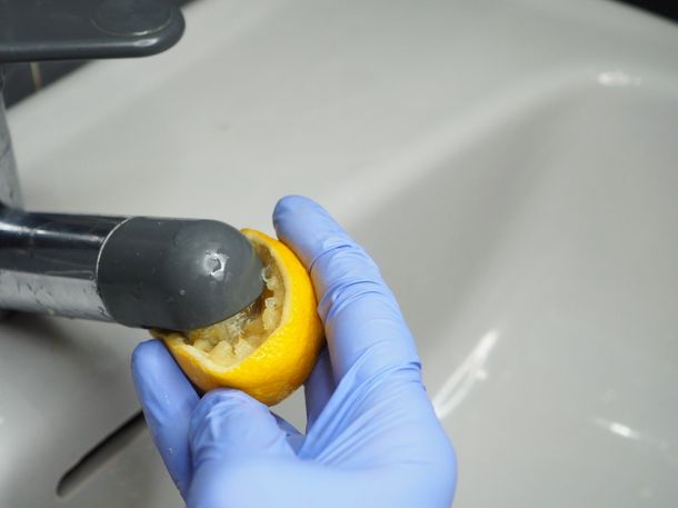 A person wearing blue gloves is holding a lemon in front of a sink faucet.