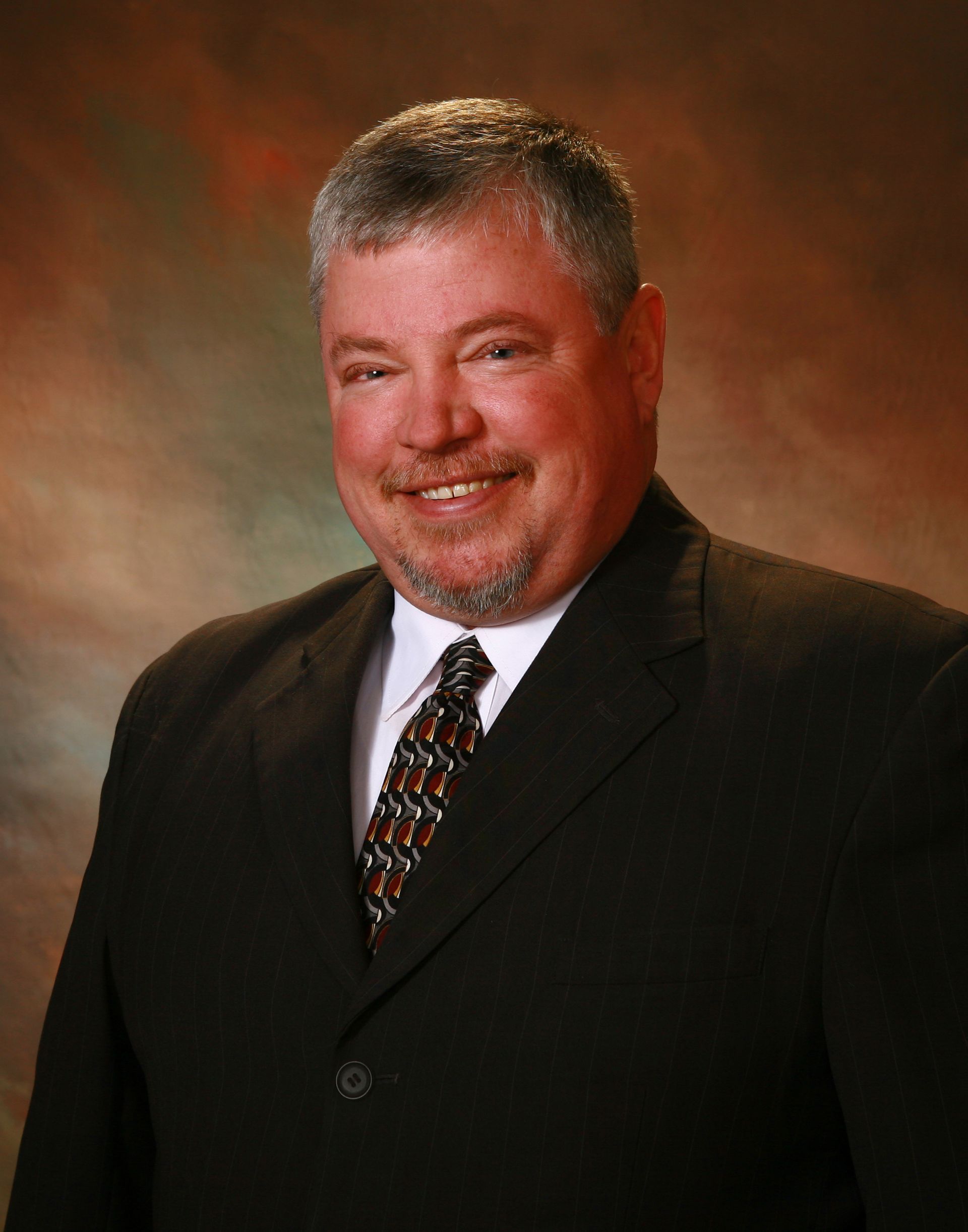 Man in a dark suit smiles, with a patterned tie against a blended background.
