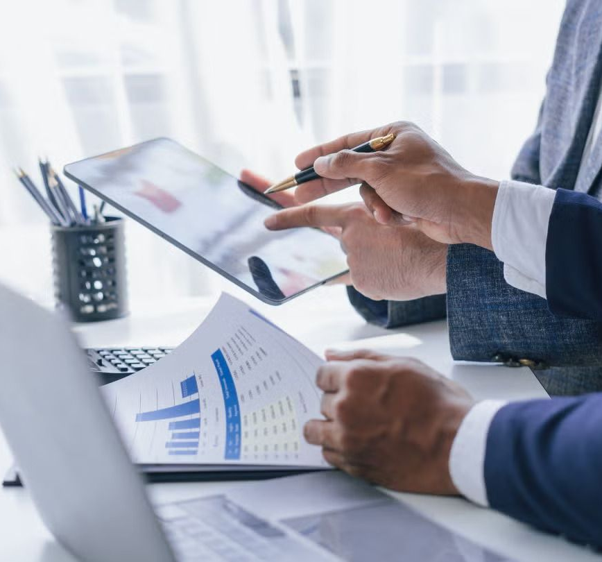 Two people in suits analyzing data on a tablet and printed charts at a desk.