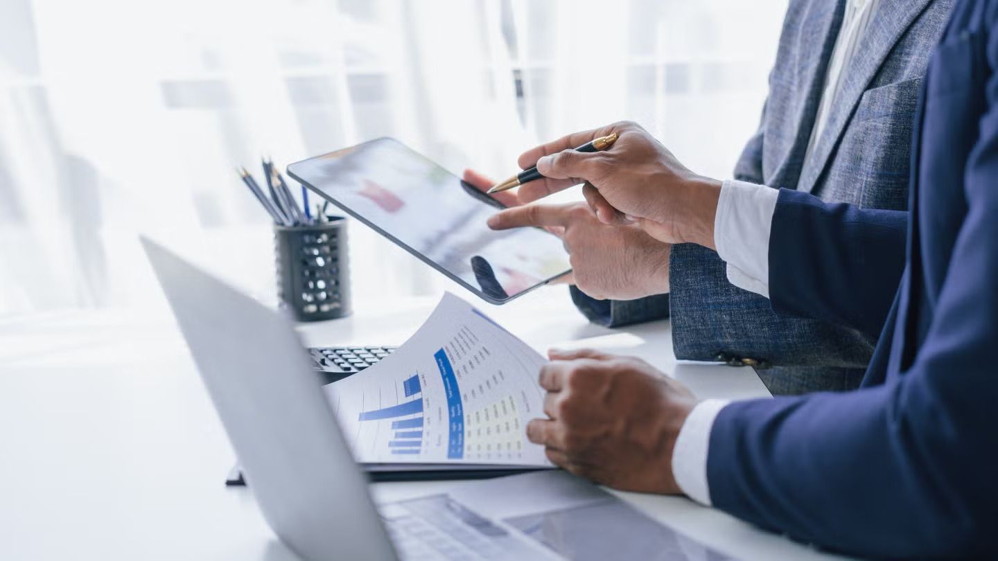 Two people in suits analyzing financial data on a tablet and printed reports.