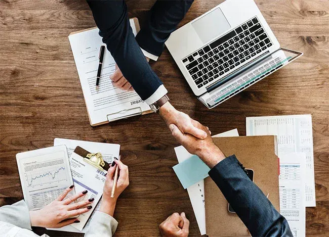 Two people in suits shaking hands over a wooden table with documents, a laptop, and a pen.