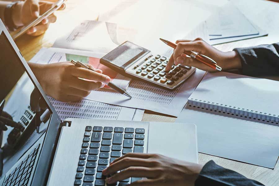 Hands using a laptop, calculator, and tablet, reviewing documents, likely financial analysis or accounting.