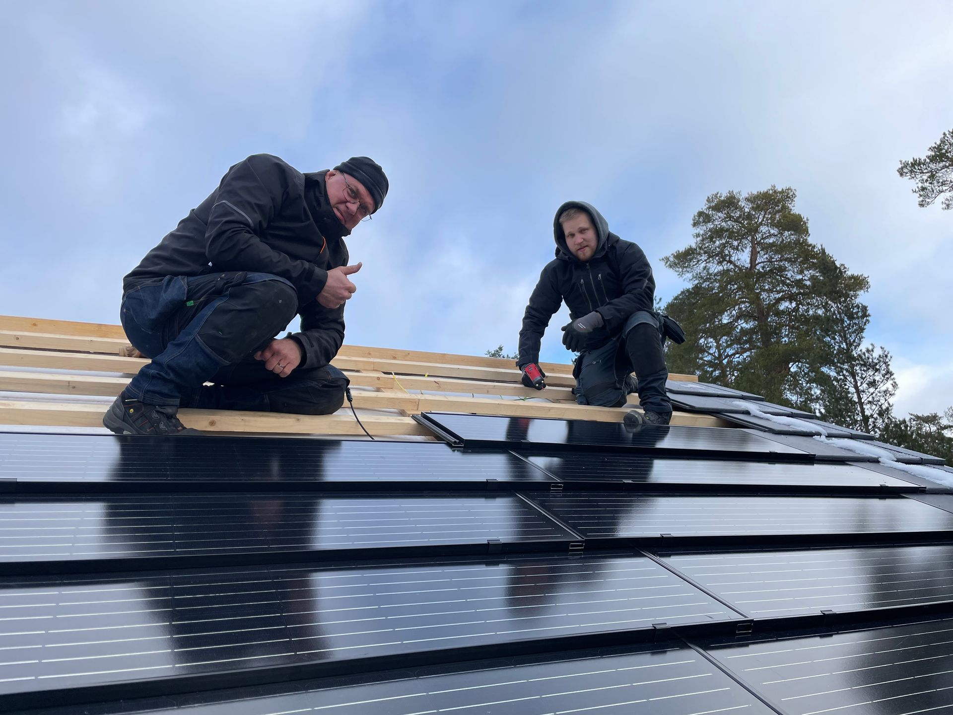 Two workers installing solar panels on a roof, thumbs-up and smiling, with snow present.
