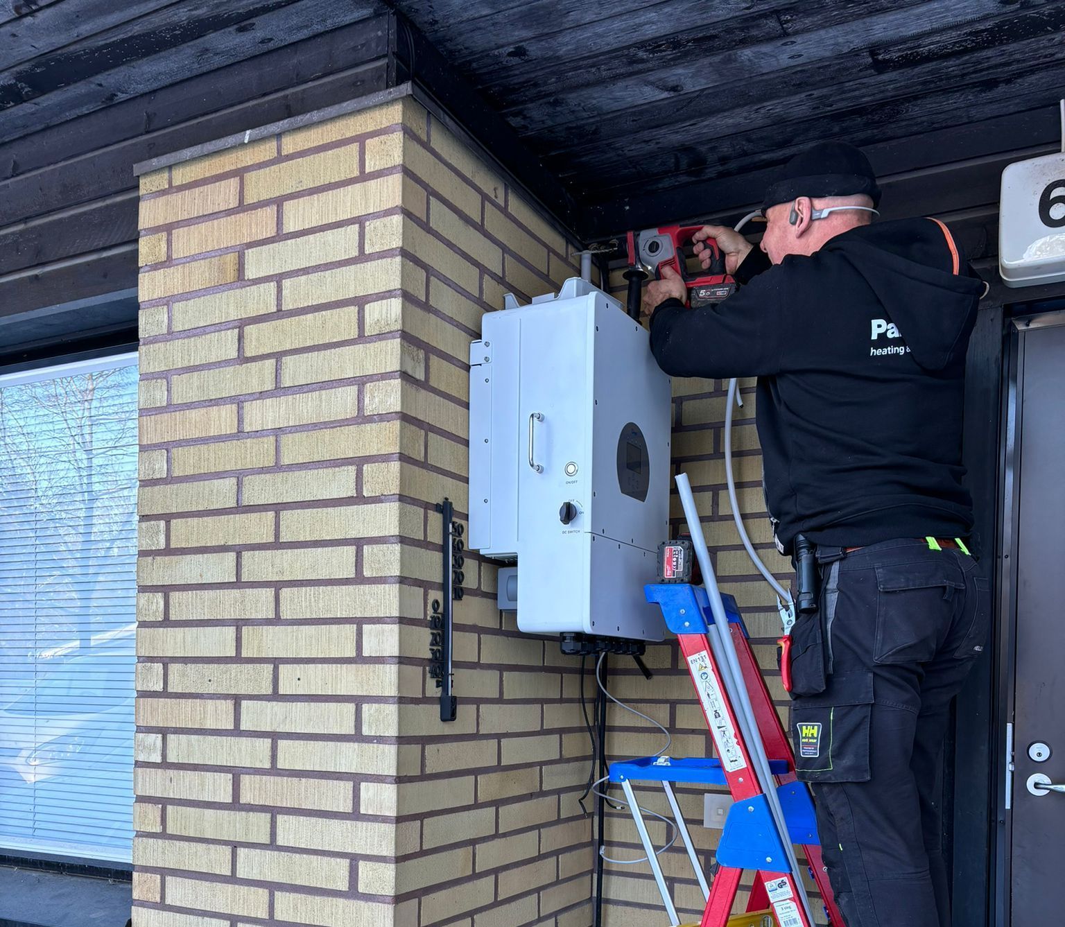 Man in black hoodie installing electrical equipment on a brick building with a ladder.