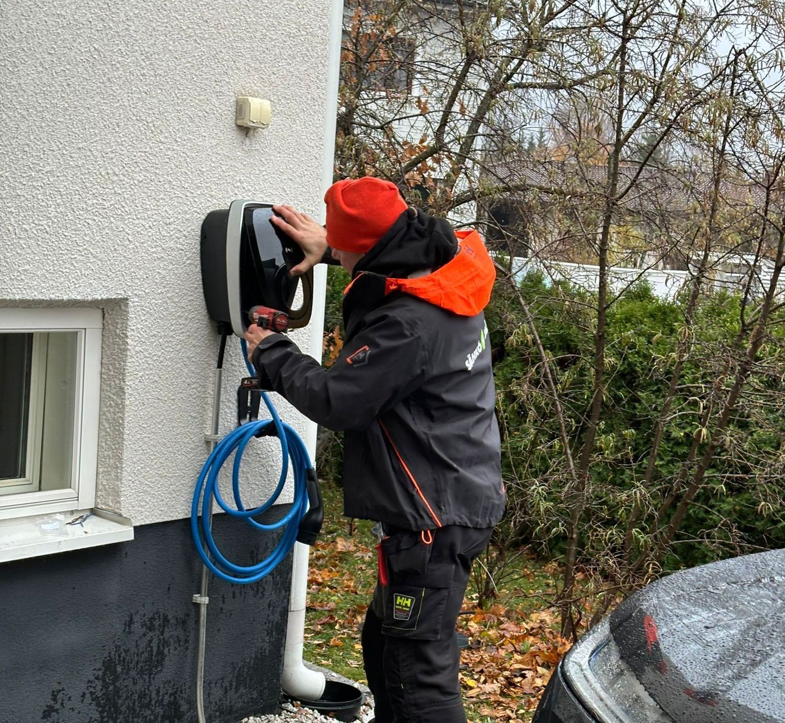 Person in orange hat installing a black EV charger on a building's exterior wall. Blue charging cable is visible.