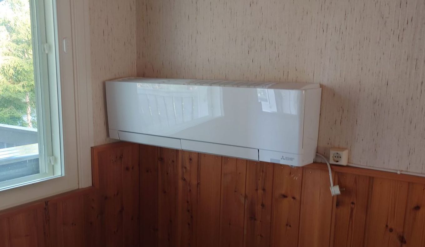 White air conditioning unit mounted on a wood-paneled wall, beside a window.
