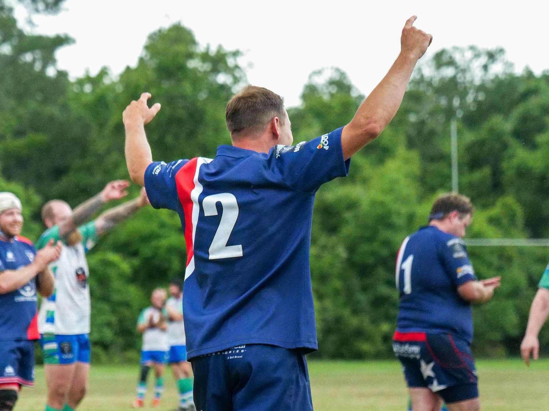 A man in a blue shirt with the number 12 on it is standing on a field with his arms in the air.