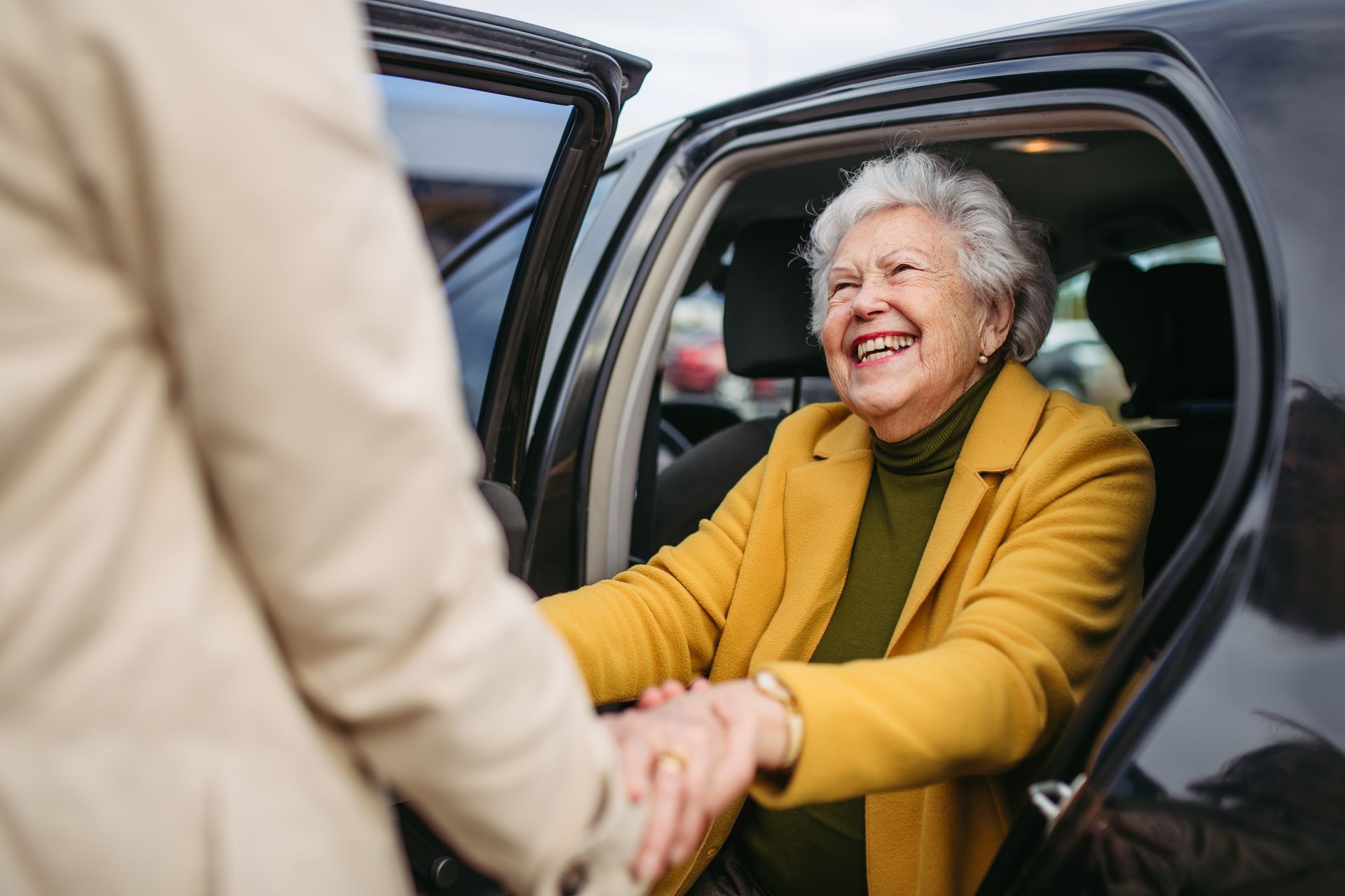 Person assisting an elderly person into a car. The woman is smiling, wearing a yellow jacket. Person assisting an elderly person into a car. The woman is smiling, wearing a yellow jacket.