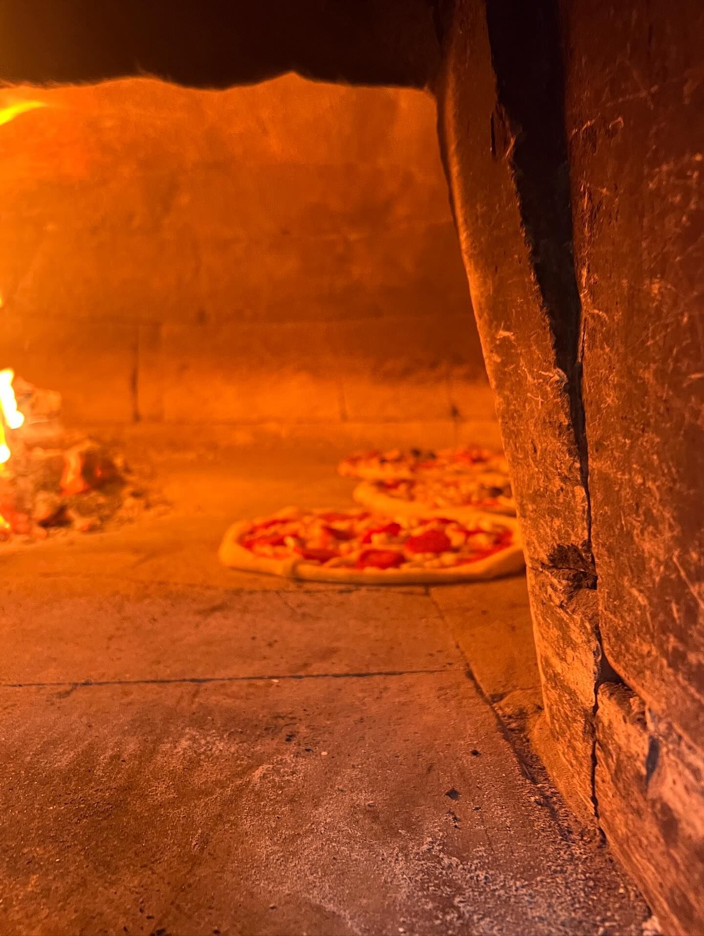 Pizze cotte nel forno a legna; fiamme e caldo bagliore arancione.
