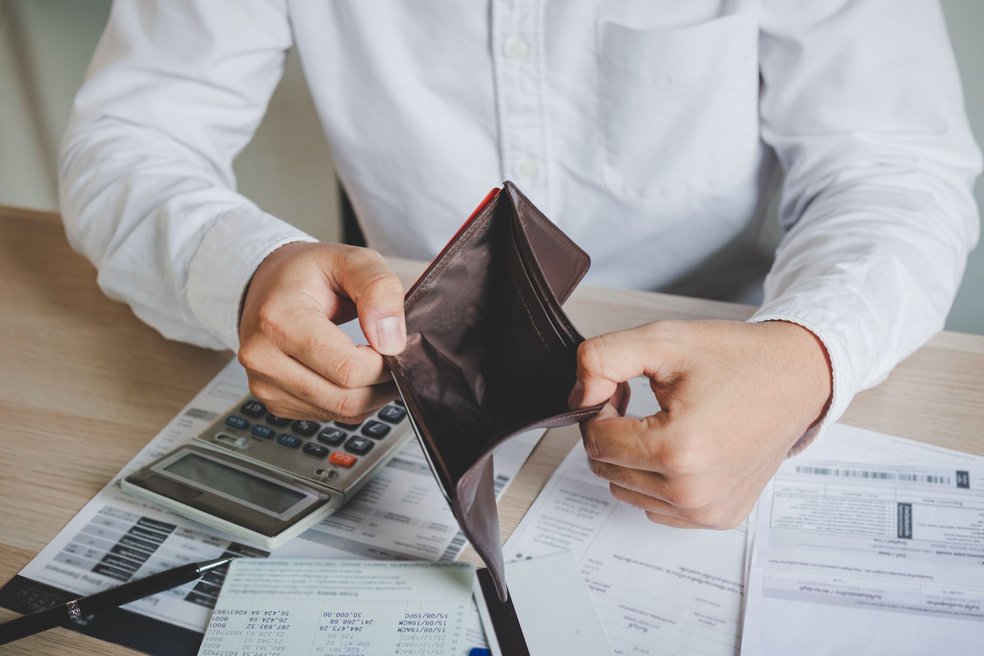A man is sitting at a table holding an empty wallet and a calculator.