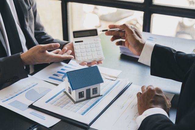 Two men are sitting at a table with a model house and a calculator.