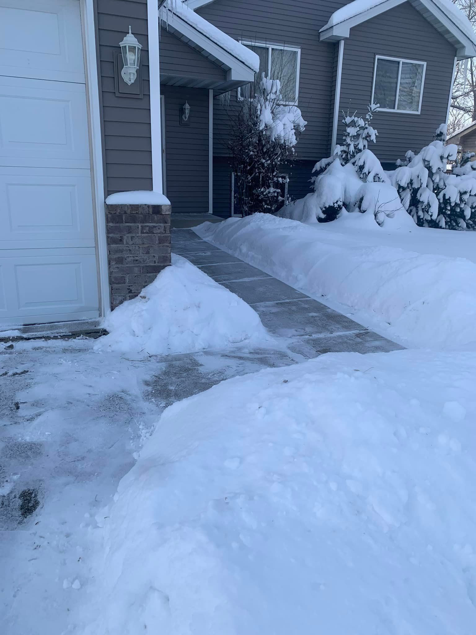 Snow-covered walkway to a house entrance, with a driveway and garage.