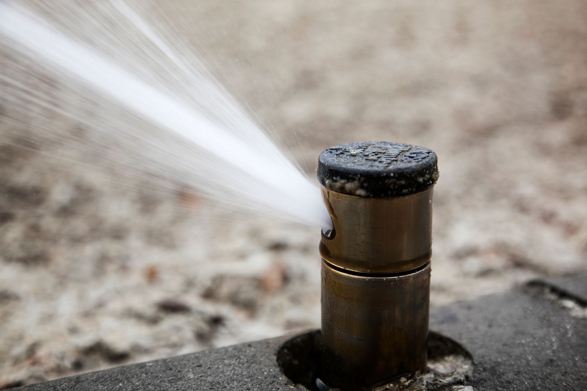 A brass lawn sprinkler head spraying a steady stream of water onto a concrete surface.