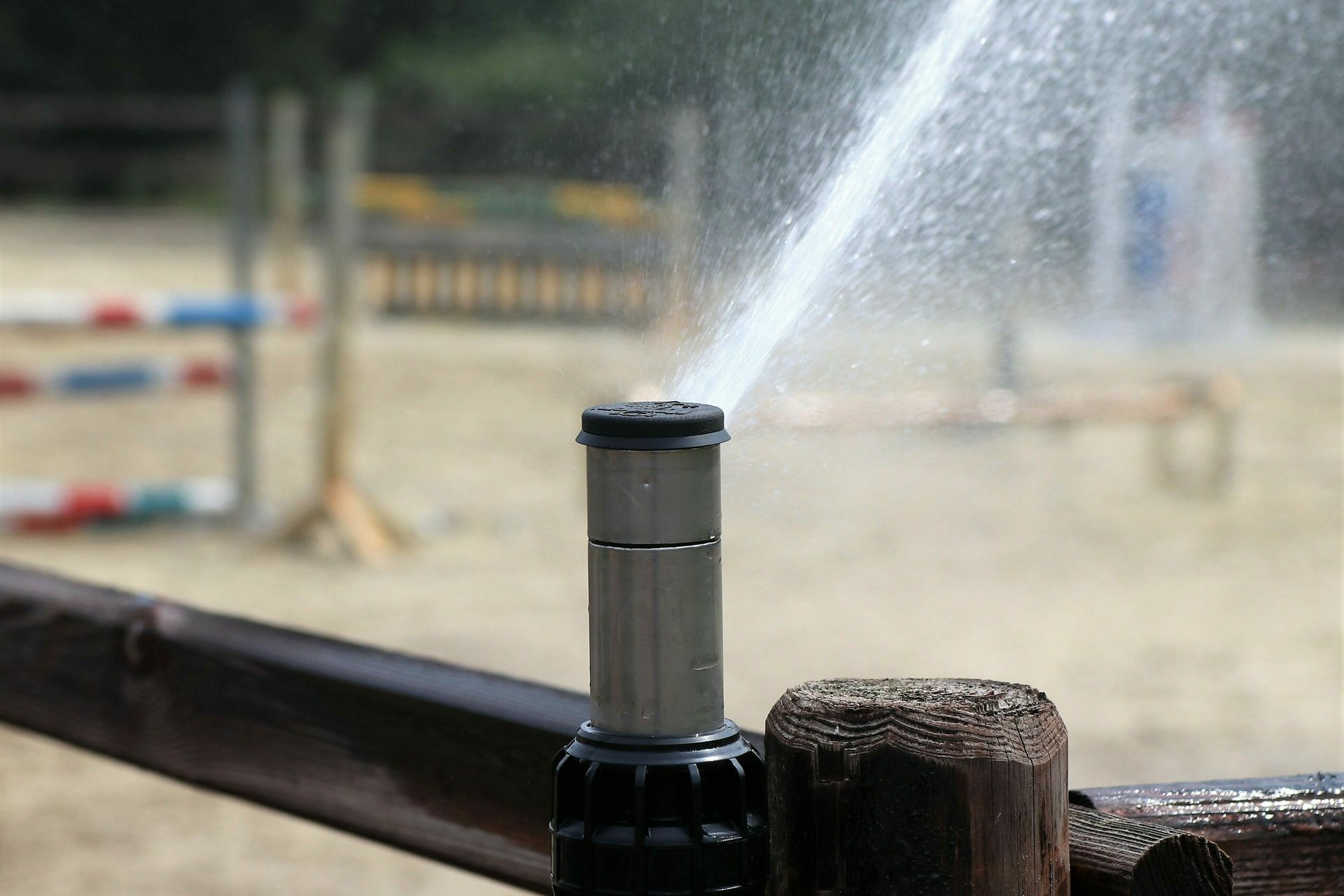 A sprinkler head spraying a jet of water over a sandy equestrian arena with training jumps in the background.