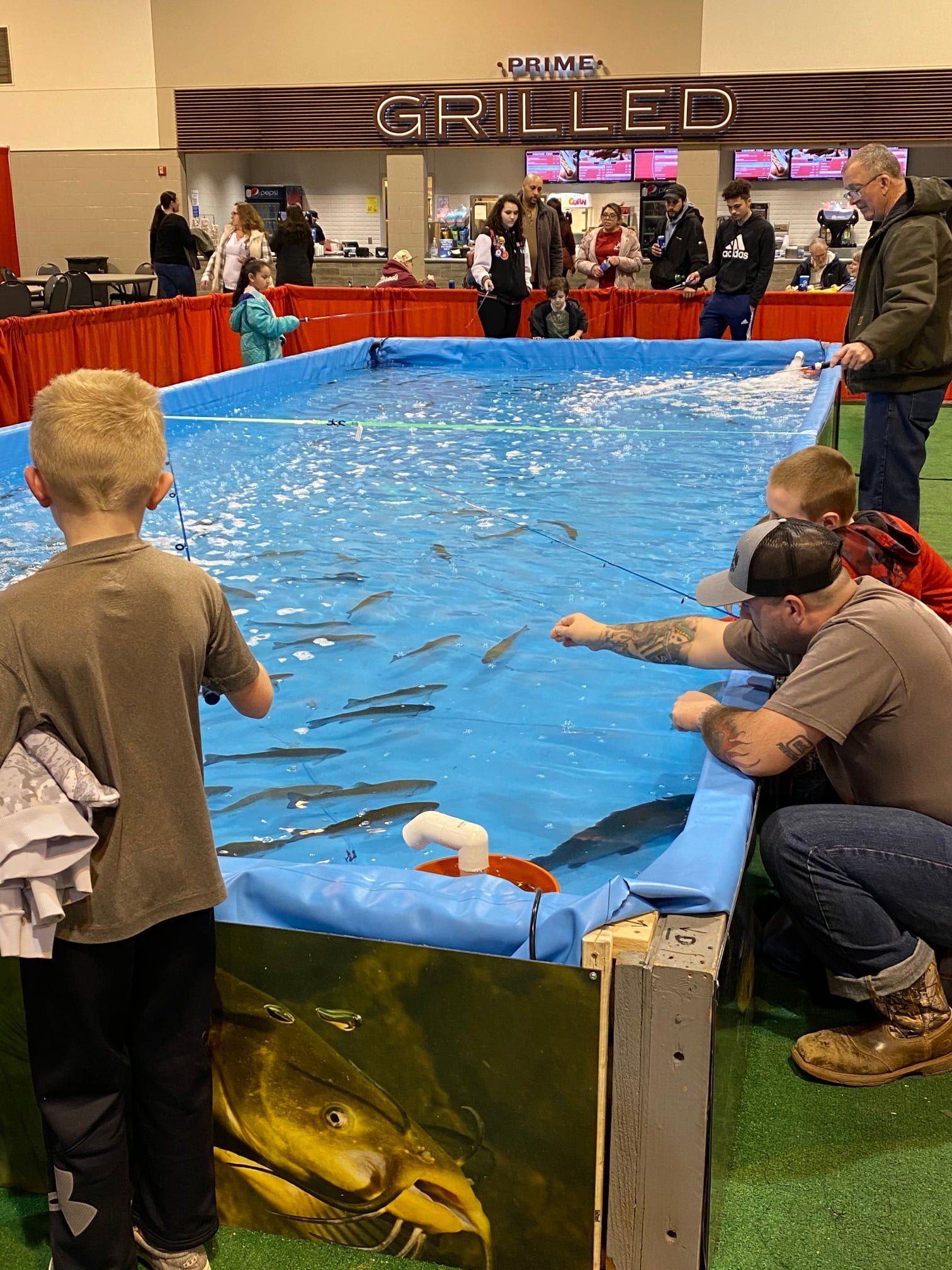 Children fishing in a pool, surrounded by adults. Indoor event with a "Grilled" sign in the background.