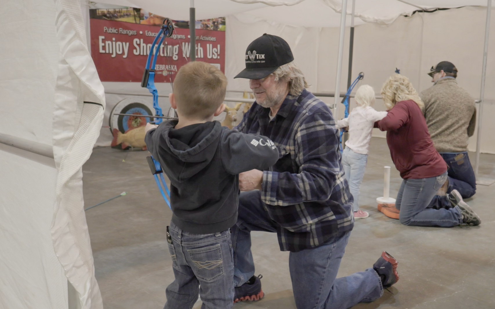 Man helping child shoot bow and arrow at an archery range. Others watching in a tent.