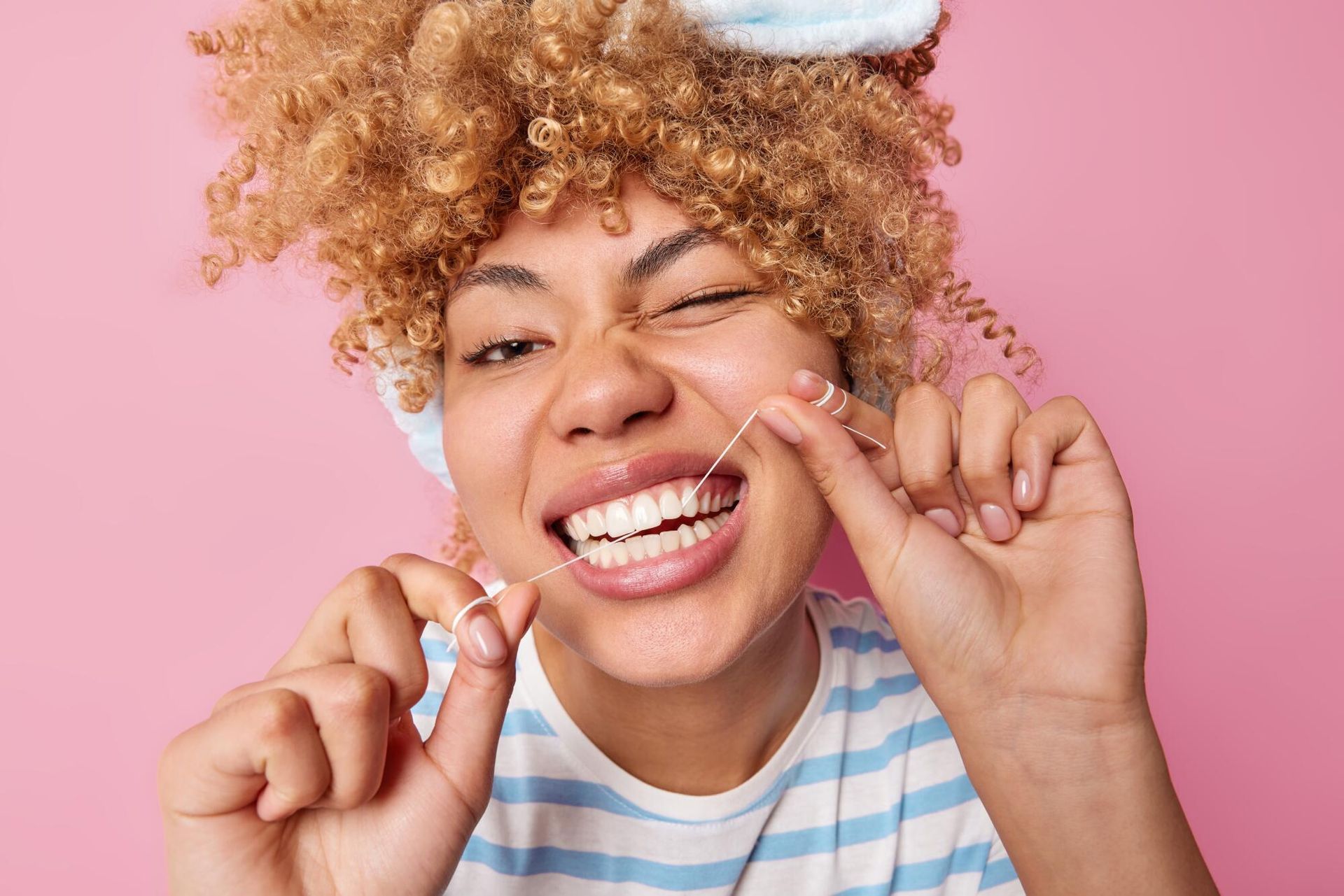 Woman flossing teeth, winking, against a pink background. She wears a headband and striped shirt.