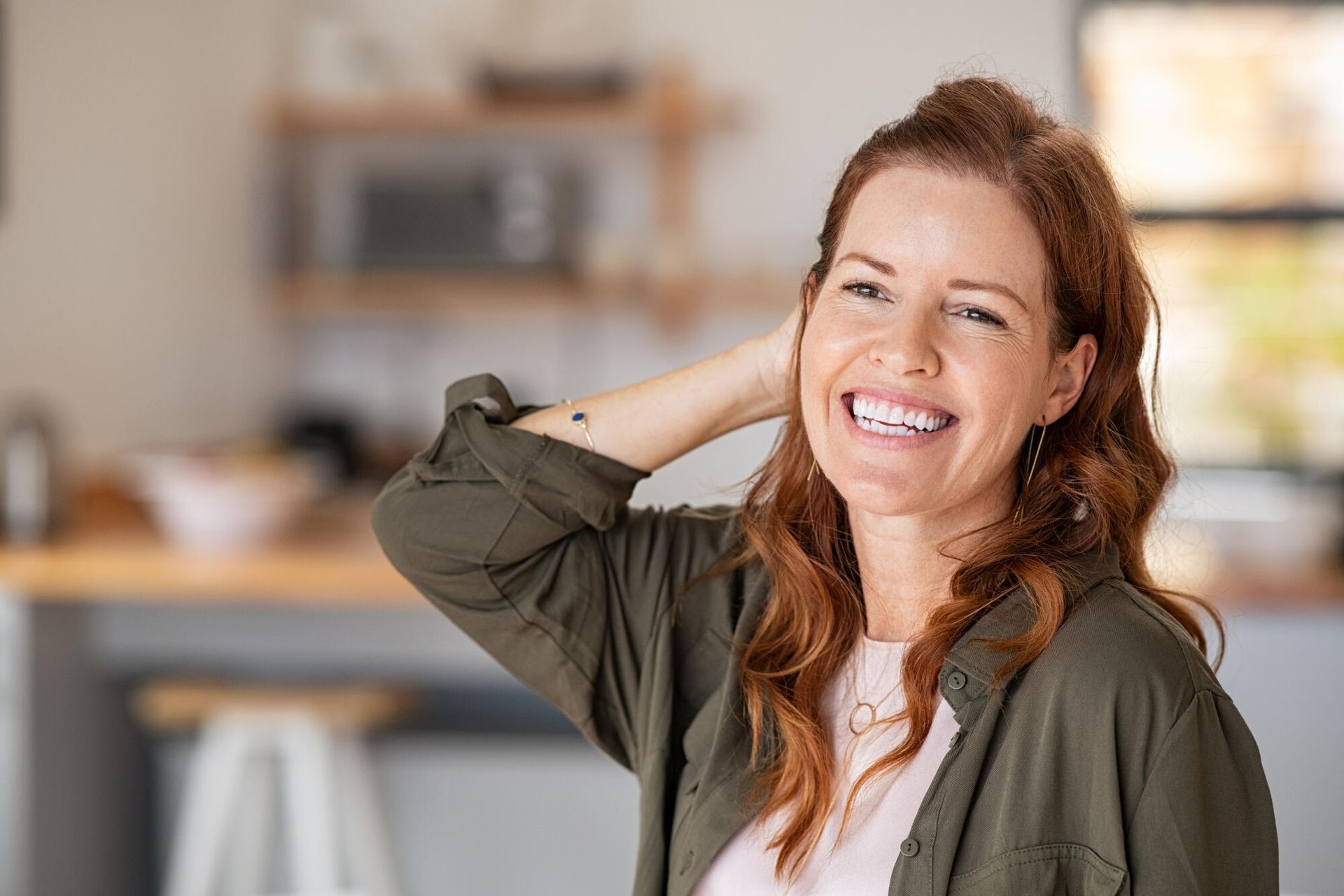Woman with red hair smiles, hand in hair, in a kitchen with a microwave, counter and stool.