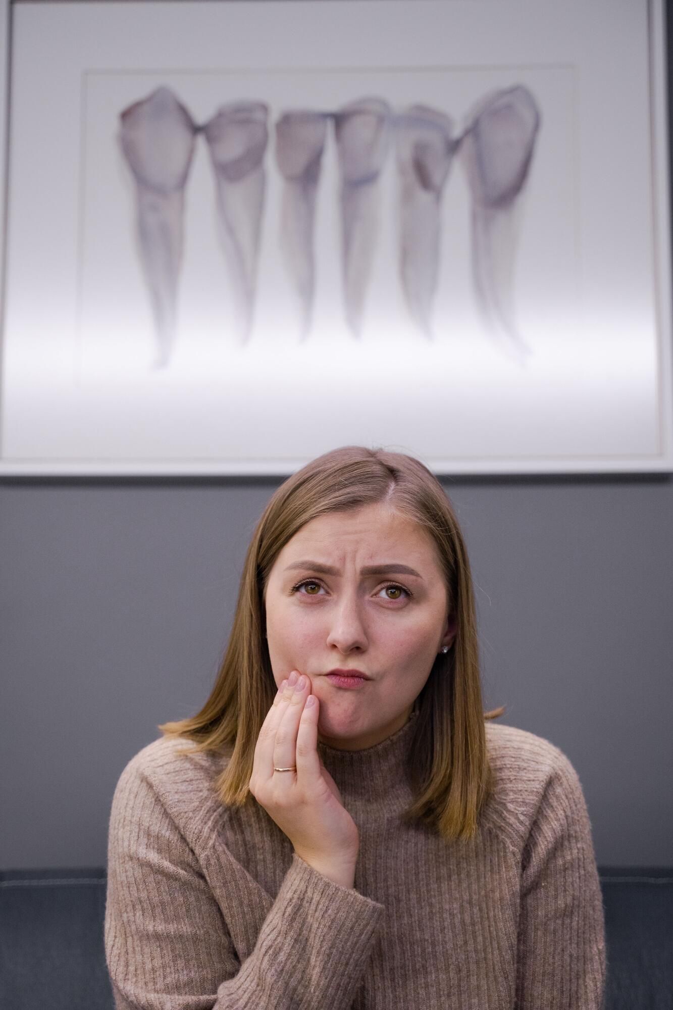 Woman sitting indoors, holding her cheek, with a pained expression and a dental diagram on the wall behind her.