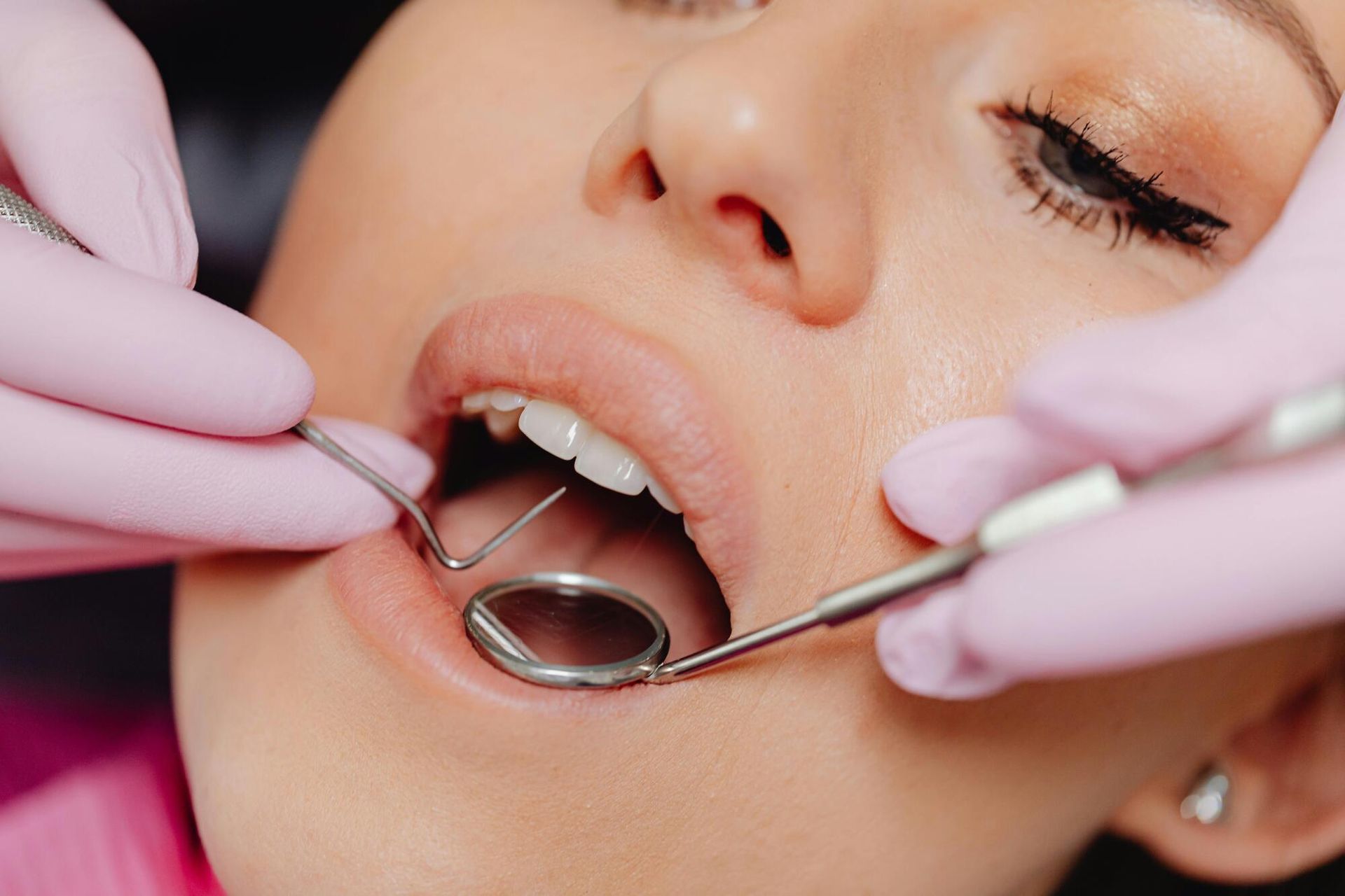 A dentist wearing pink gloves uses a mirror and a probe to examine a patient's teeth during a dental checkup.