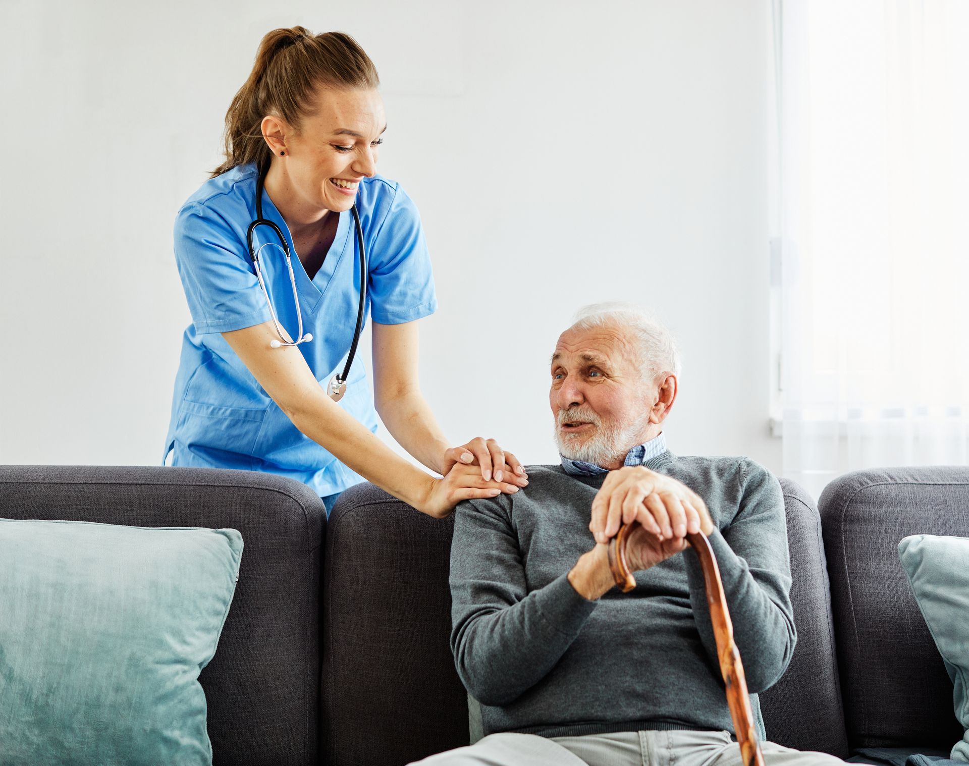 Nurse smiles, placing a hand on the shoulder of an elderly man sitting on a sofa, holding a cane.