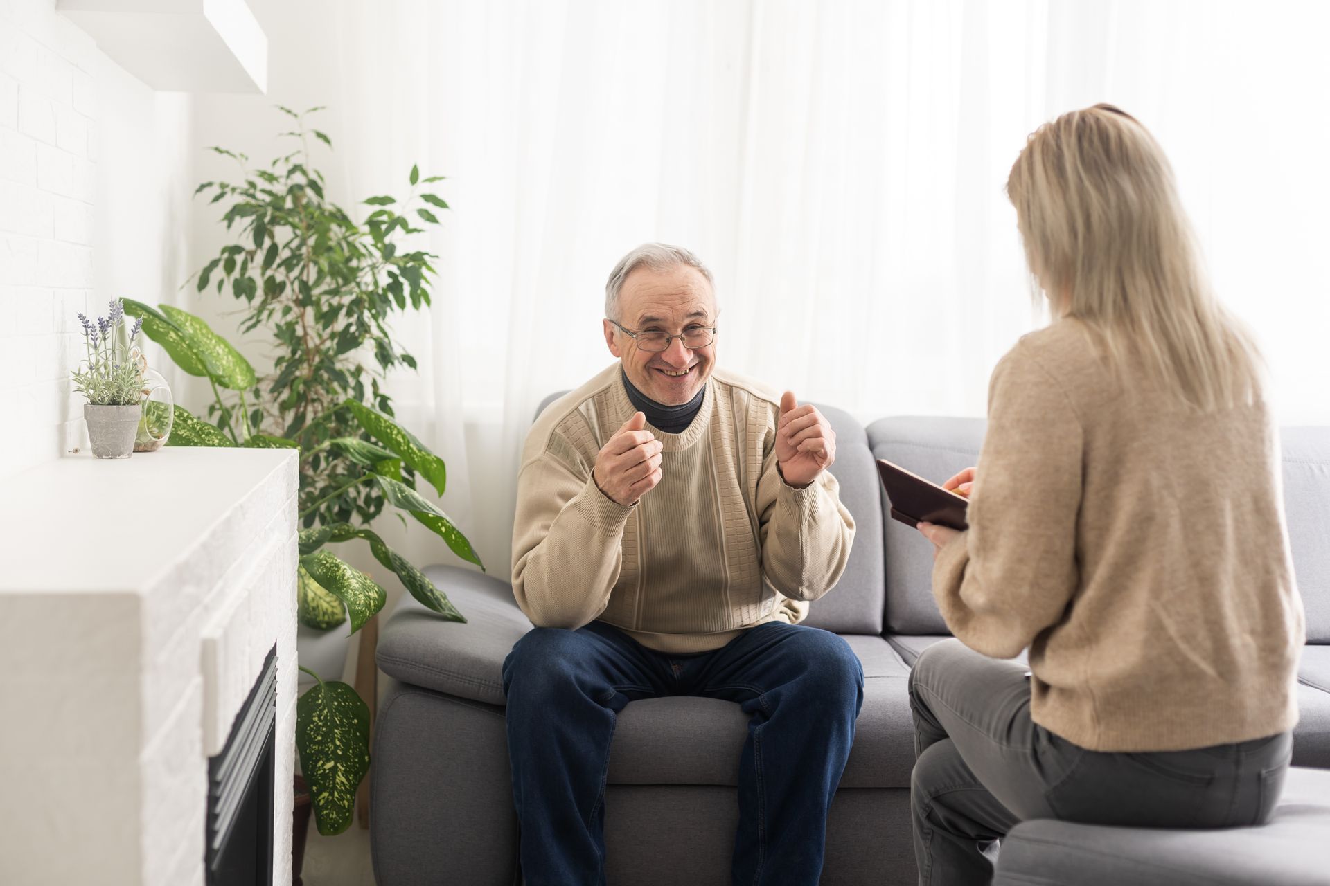 Elderly man smiles, gestures while a woman plays guitar on a couch indoors.