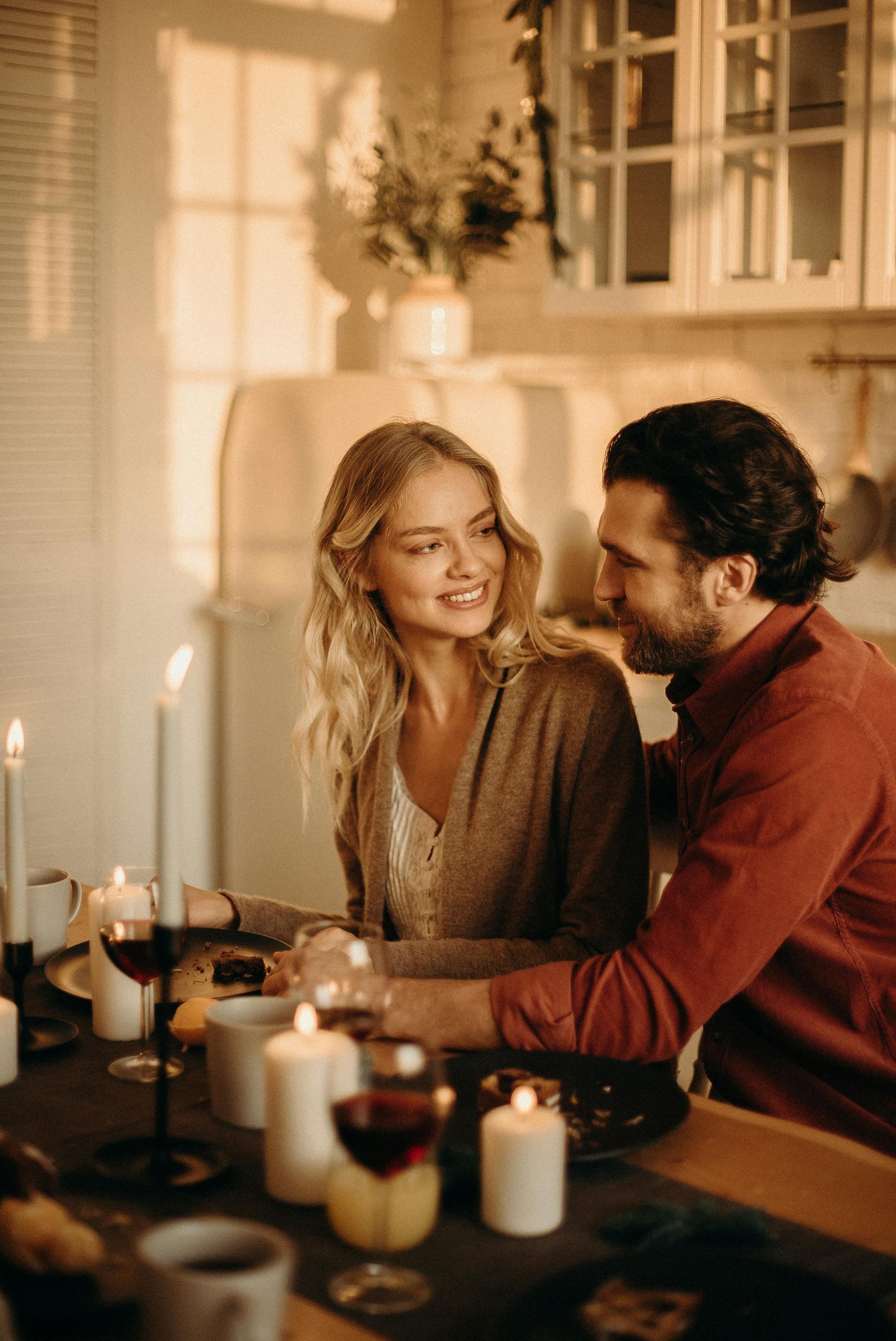 Couple smiling at each other during a candlelit dinner. Blonde woman, bearded man. Cozy, indoor setting.