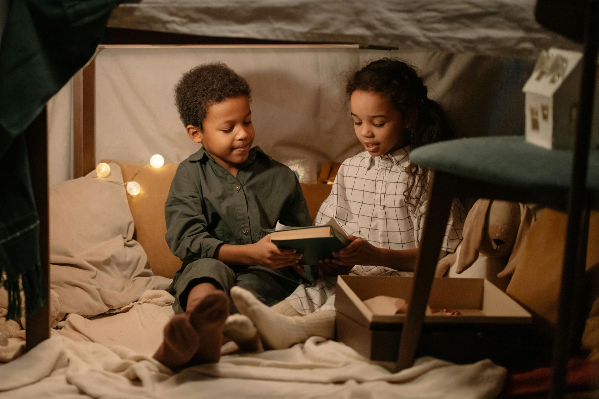 Two children share a book inside a blanket fort, lit by fairy lights.