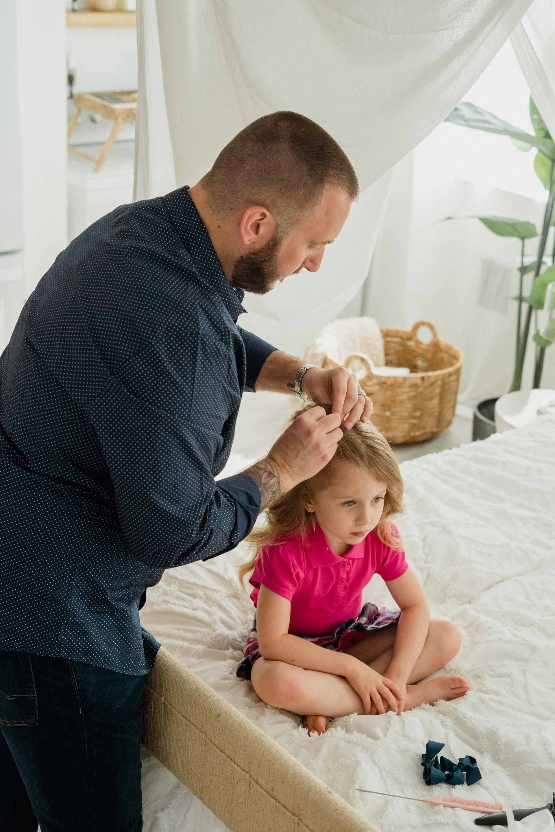 Man fixing girl's hair on a bed. Girl in pink shirt looks down; they're in a light-filled room.