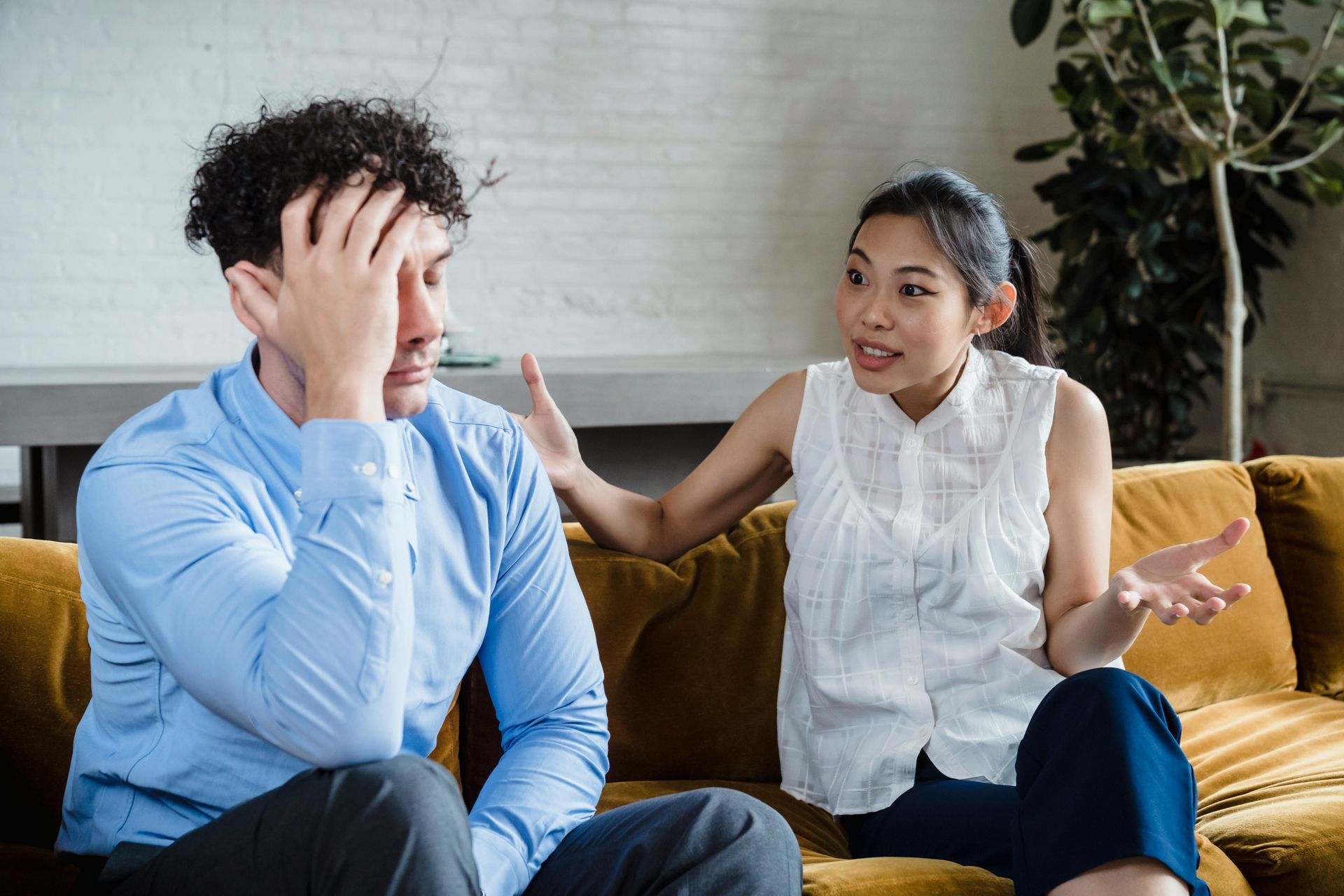 Woman gesturing, arguing with a man on a sofa. Man has hand on his head.