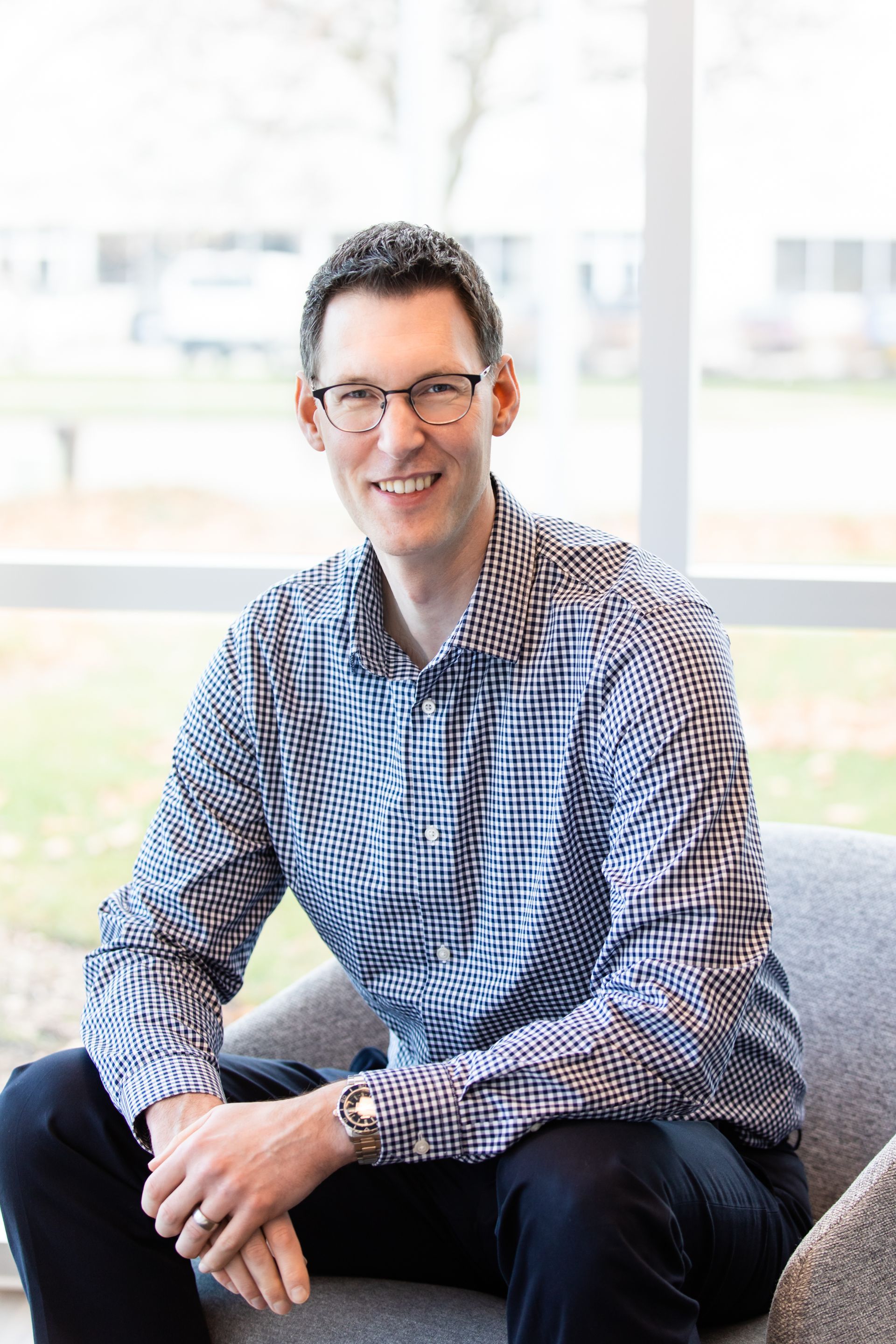 A man wearing glasses is sitting on a couch in front of a window.