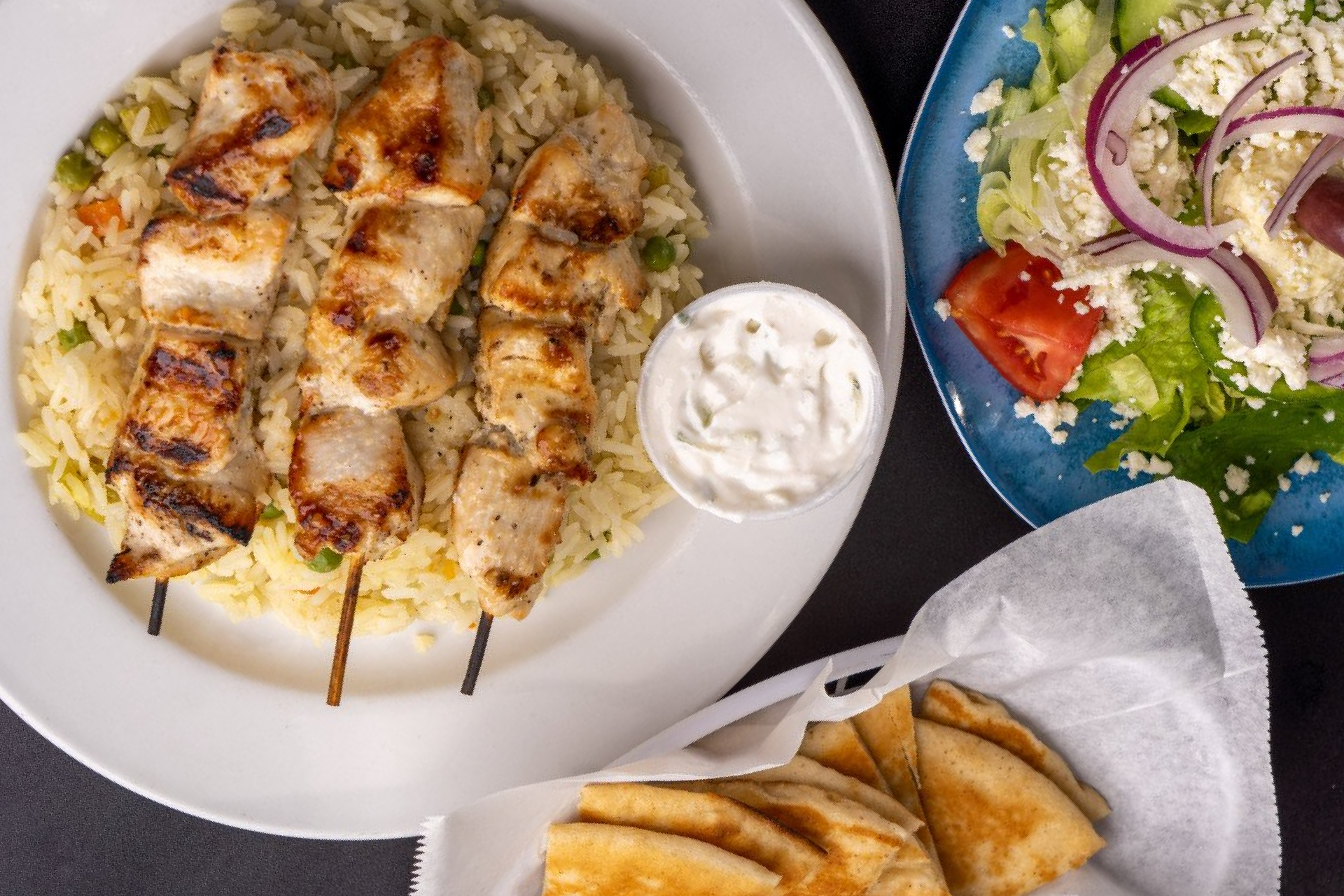 A white plate topped with chicken skewers and rice next to a salad and pita bread.