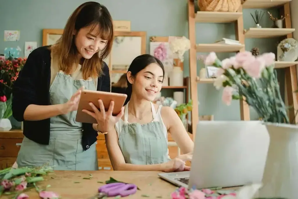 Two women in aprons working in a flower shop, one on a laptop, the other with a tablet, flowers on the table.