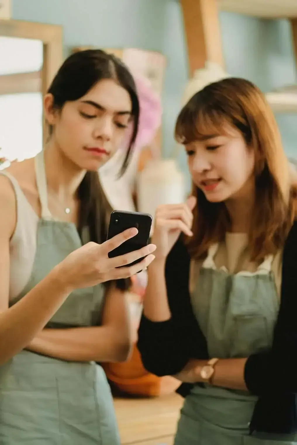 Two women in aprons look at a phone, one pointing, in a shop setting.