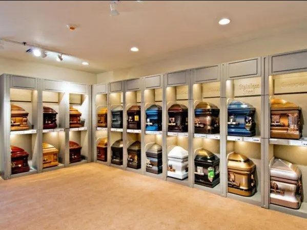 Casket showroom with various colored coffins on display, lit by overhead lights, beige walls and flooring.
