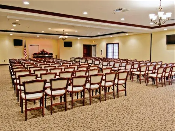 Rows of chairs fill a beige-carpeted room, facing a stage with an American flag, likely a meeting hall.