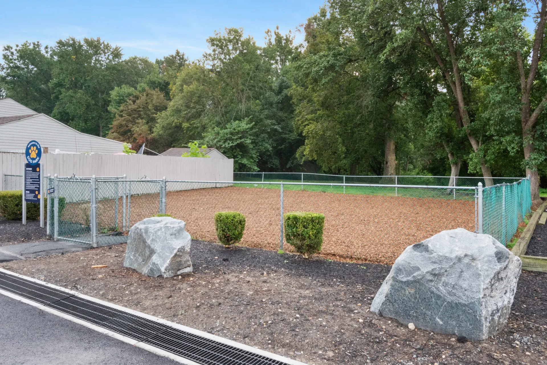 Fenced dog park area with gravel surface, rocks, and surrounding trees.
