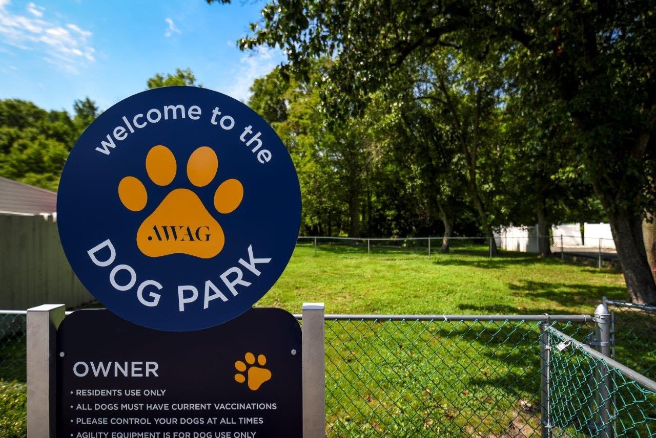 Circular blue dog-park sign with a yellow paw print, beside a chain-link fence and grassy area.