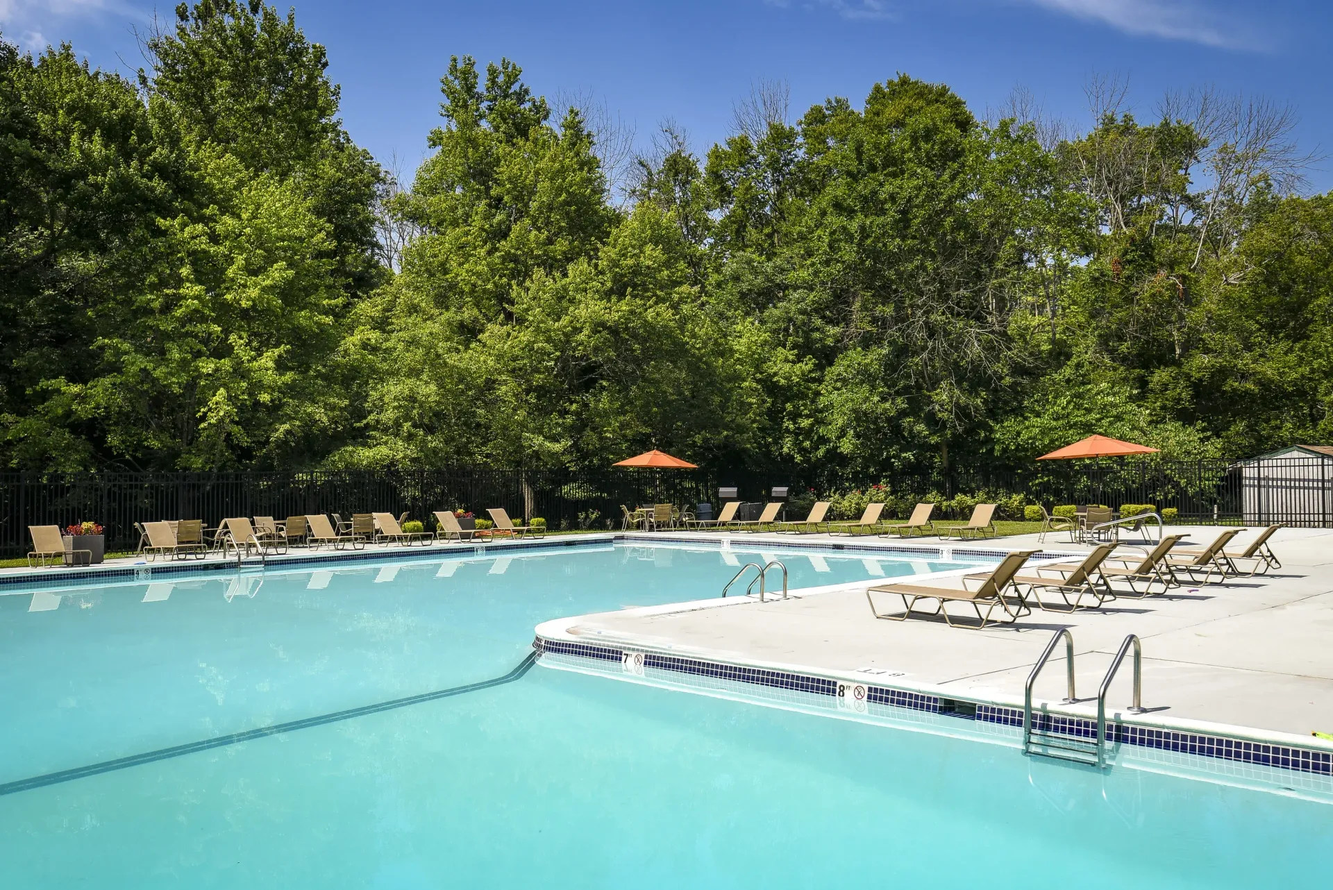 Outdoor community pool with lounge chairs and orange umbrellas, framed by trees.