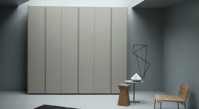 Una stanza minimalista caratterizzata da un grande armadio beige a tutta altezza, un tavolino rotondo, uno sgabello geometrico e una sedia.