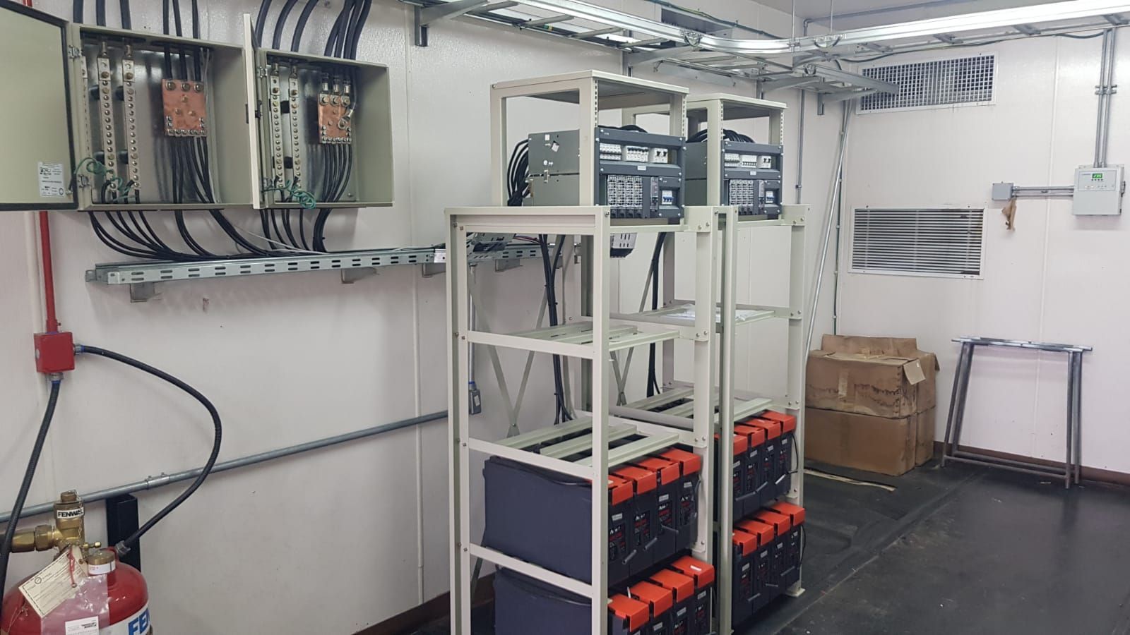 Sala de control eléctrico industrial con bastidores de equipos, paneles de cableado y unidades de baterías en estanterías.