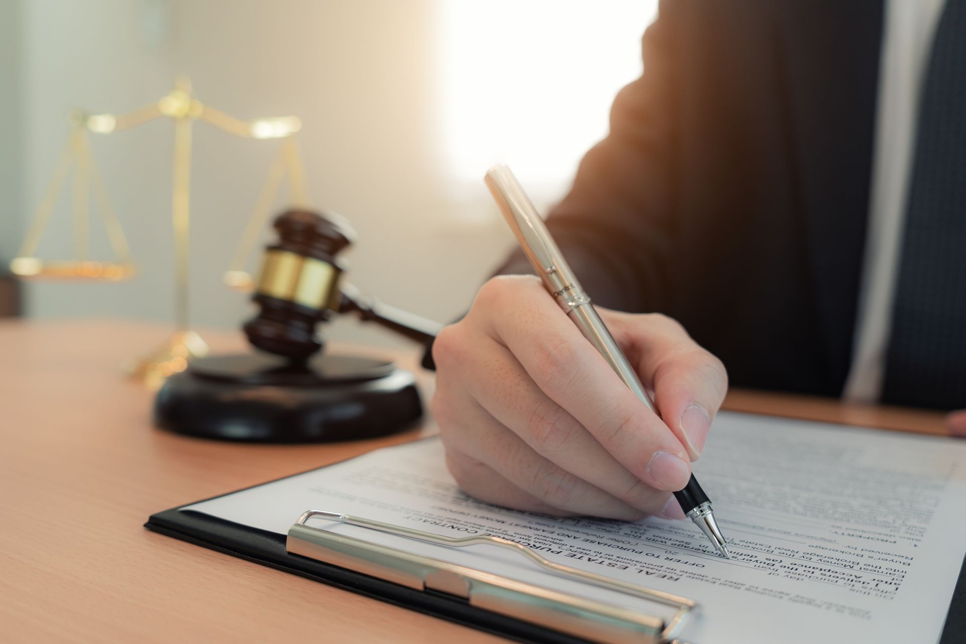 Person in suit writing on a legal document with a gavel and scales in the background.