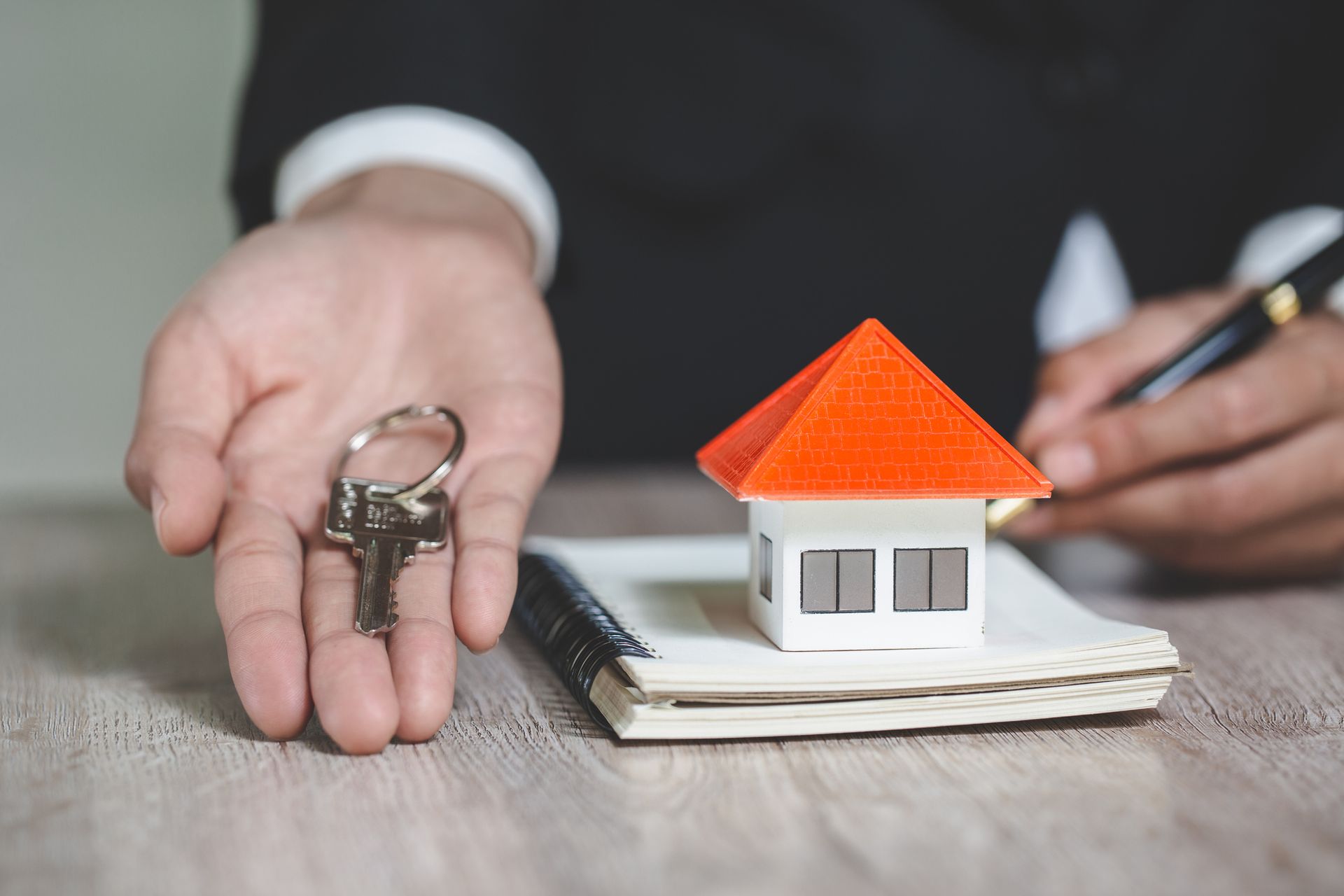 Person holding keys and a tiny house on top of a notebook, signing a document.