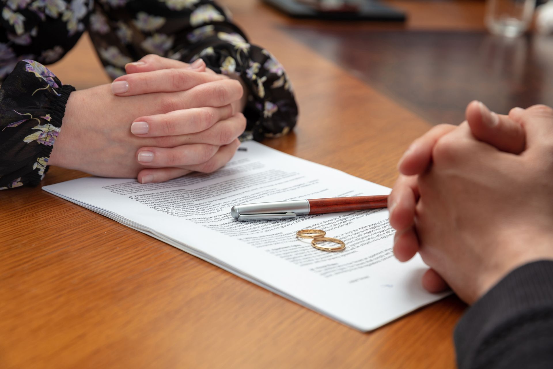 Hands clasped over a document on a wooden table, wedding ring, pen; likely a divorce negotiation.