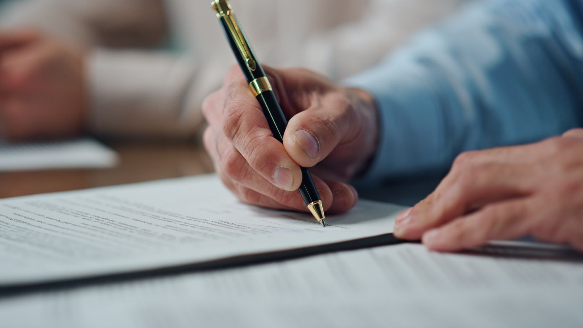 A person's hand signing a document with a black and gold pen, light blue shirt.